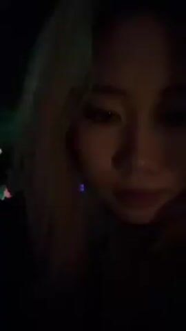 A young woman with light blonde or brown hair looks into the camera. She is in a dark environment with shifting blue and purple lighting, reminiscent of a nightclub or concert venue. Blurred figures and lighting fixtures can be seen in the background. The woman has fair skin and dark eyes. There isn’t much activity in the video.