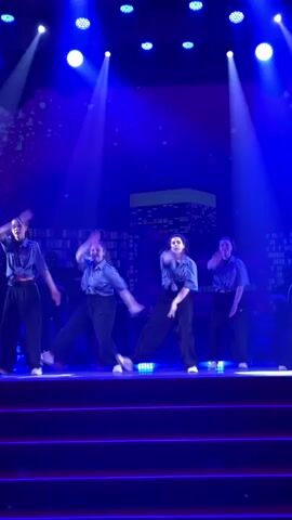 On a stage with a backdrop of a city skyline and bathed in blue light, a group of young women perform a choreographed dance routine. They are dressed in matching outfits: light blue, button-down shirts and dark, wide-legged pants. Their movements are synchronized and energetic as they step, kick, and swing their arms in unison. The stage has a red surface with neon lighting along the edges. The atmosphere is vibrant with the lighting and music creating an energetic performance.
