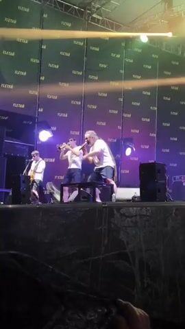 This is a video clip of a live musical performance on an outdoor stage. Three men are visible on stage. The man on the left is playing a guitar. The man in the center is playing a trumpet. The man on the right, who has long dreadlocks, is holding a microphone and appears to be singing. He is also playing a saxophone. The stage is set up with a backdrop of fabric panels covered in the repeated text "YLETAЙ". There are several large speakers on either side of the stage, and spotlights are illuminating the performers and the stage. The lighting suggests it is evening or nighttime. There are some indications of rain or moisture on the stage in the foreground. The overall scene suggests a festival or concert event.
