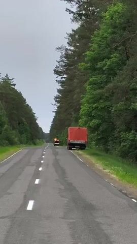 A dashcam video shows a road lined with trees. A red-covered truck is parked on the right side of the road. Further down the road, another vehicle with flashing lights is visible. The road is wet, suggesting recent rain. The sky is overcast.