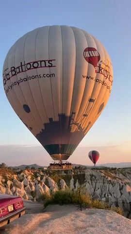 A large, white hot air balloon, bearing the "Kaya Balloons" branding and website ("www.kayaballoons.com"), dominates the frame, either preparing for or having just completed a launch in Göreme, Turkey. Passengers are visible in its wicker basket, enjoying the expansive views. The balloon is adorned with a red company logo, a silhouette of a city or castle at its base, and further text on its right side, including "Kapitalo Kaya Balloons" and a partial web address likely related to Kapadokya.

In the background, a smaller, striped hot air balloon (red and white) drifts gracefully against the clear sky, indicative of other flights taking place. The scene is bathed in the warm, golden light of either sunrise or sunset, casting long shadows and highlighting the unique geological formations characteristic of Cappadocia—jagged rock pillars and valleys with sparse green vegetation. The sky is a gradient of light blue near the horizon, deepening to a richer blue overhead.

In the lower left foreground, the rear of a vintage-style red car is partially visible, adding a pop of color and a classic aesthetic to the rugged landscape. The overall setting is one of serene beauty and adventure, capturing the iconic hot air balloon experience in this famous Turkish region. The weather appears perfectly calm and clear for ballooning.