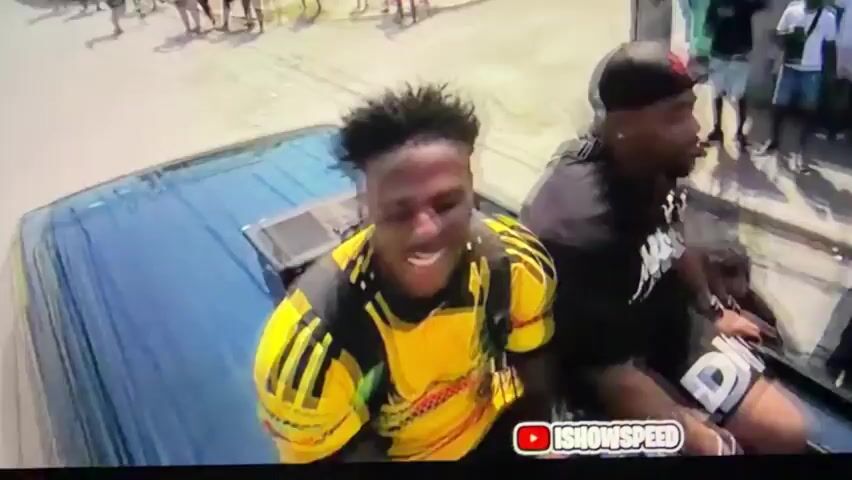 The video shows two Black men riding on the roof of a dark blue SUV. The man on the left is wearing a yellow Jamaican soccer jersey and a black backpack. The man on the right is wearing a black t-shirt that says "Jamaica" and a black baseball cap with a red logo on it. The car is moving along a street in what appears to be a Jamaican neighborhood. People are standing on the sidewalks, and some are watching the car go by. A billboard with a drawing of a lion can be seen. The words "ISHOWSPEED" are at the bottom of the screen.