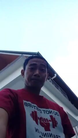 A man in a red t-shirt is filmed from the chest up. The t-shirt has a white design with text and a graphic of a weightlifter's fist and a dumbbell. The man is looking at the camera and smiling slightly. He is outdoors, with a modern building visible in the background. The building has white walls, a dark roof, and a balcony. The video is shot from a low angle and appears to be a selfie video.
