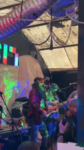 A band is performing live in a venue with unconventional decor. The singer/guitarist is in the foreground, dressed in a red plaid shirt and a headband, with long dark hair and a beard.  He is playing an electric guitar and singing into a microphone. Behind him are other band members, including another guitarist and a bassist. The setting is dimly lit, with colorful lights flashing and a projector displaying abstract patterns on the wall. The ceiling has exposed pipes and ductwork, and the overall atmosphere is somewhat grungy.