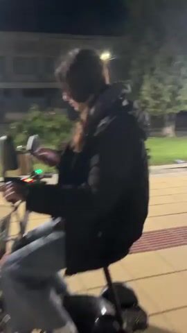 A young woman with long dark hair, wearing a black puffer jacket and blue jeans, is seen outdoors at night in what appears to be a park or pedestrian area. Streetlights illuminate the scene, with trees, benches, and buildings visible in the background.

At the beginning of the video, she is looking down at a smartphone, which she holds in her left hand. The screen of the phone is briefly visible. She is seated on an electric scooter or bicycle, with a small basket attached to the front handlebars. A voice off-camera asks, "Hello, I have one question. What do you hate?"

The woman looks up, makes eye contact with the camera, and smiles briefly before replying, "Men." She then chuckles and quickly starts to ride the electric scooter away from the camera, moving from left to right across the frame. Her laughter continues as she accelerates. The location is Lozarevo, Bulgaria.