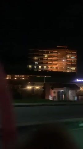 The video is a nighttime shot of a hotel. The hotel is a tall, multi-story building with many windows lit from within. In front of the main building is a lower structure with an illuminated sign that reads "HOTEL PRESIDENTIAL." There is a street in the foreground, and a car passes by. The overall lighting suggests it is dark outside.