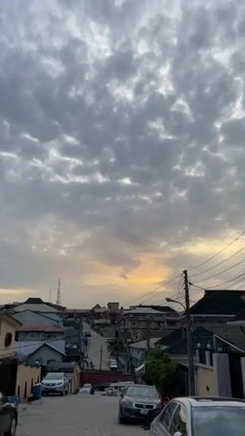 The video captures a street view in a residential area under an overcast sky. The sky is filled with grey and white clouds, with a hint of golden light peeking through near the horizon. The neighborhood features a variety of houses and buildings with different architectural styles. Utility poles and wires crisscross the sky above the rooftops. Cars are parked along the side of the road, and a person is visible near a silver vehicle. The road slopes downwards, and the overall scene suggests either dawn or dusk.
