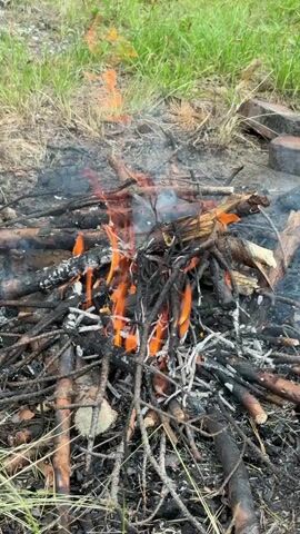 The video shows a close-up view of a campfire burning in a grassy area. The fire is composed of numerous small, dry branches and twigs, with some larger pieces of wood. Flames are actively burning, with orange and yellow hues visible. Smoke rises from the fire, appearing as a thin, white plume that drifts to the left. The ground around the fire is covered in ash, charred wood, and dried leaves. In the background, lush green grass and vegetation are visible, suggesting an outdoor, possibly wooded, environment. The overall impression is of a small, controlled campfire.