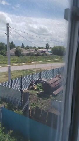 The video shows an outdoor scene on a cloudy day. In the foreground, a metal fence is visible, and behind it, there is a pile of debris including what appears to be a large, rusty metal tank or barrel. Further back, another metal fence encloses a yard. Beyond the fences, there is a road and a grassy area with some trees and houses in the distance. A utility pole with wires is also prominent in the middle ground. The overall impression is of a somewhat neglected rural or semi-rural environment.