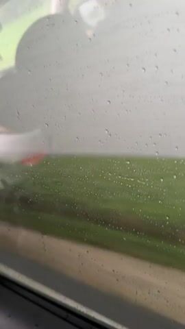 The video shows a close-up view of a window with raindrops on it. Through the window, a blurry green landscape is visible, suggesting an outdoor environment. The motion of the camera appears to be slightly shaky and possibly moving. In the foreground, on the left side of the frame, a white object with a hint of red is partially visible, touching the glass. The overall impression is that the footage was captured from inside a moving vehicle during rainy weather.