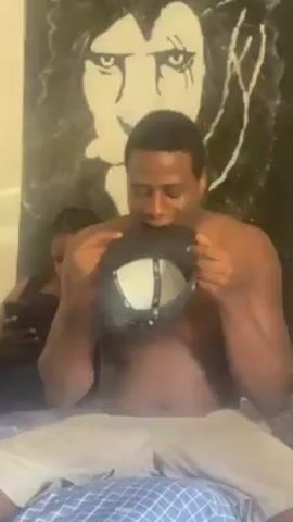 The video shows a shirtless, dark-skinned man sitting cross-legged on a bed, holding a black and white baseball cap.  He first looks down at the cap, then up at the camera. He then bounces the cap on his hand, and brings it to his mouth, touching it to his lips. The bed is covered with a light blue patterned comforter.  Behind the man is a wall with a painting of a lion.  Another person is vaguely visible in the background to the left.