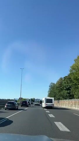 The video shows a view from inside a vehicle traveling on a highway. The sky is clear and blue with some wispy clouds. The road is asphalt with white lane markings. There is moderate traffic, with several cars and a camper van visible ahead. The vehicle appears to be in the right lane. Trees and a sound barrier line the right side of the highway. A tall pole with a light fixture stands on the left side of the road, near a traffic sign. The overall impression is a sunny day on a highway with some traffic congestion.