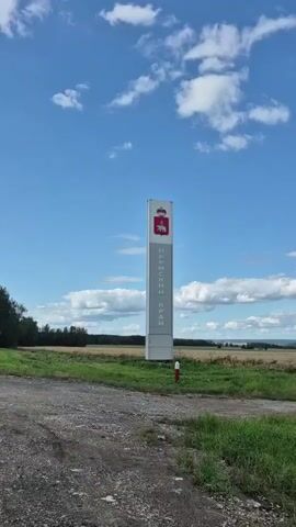 A tall, rectangular sign stands in a field under a blue sky with white clouds. The sign is white with a red crest at the top featuring a bear and crown. Vertical text in grey reads "ПЕРМСКИЙ КРАЙ". A gravel road and green grass are in the foreground.