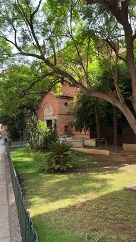 The video opens with a view of a brick building, likely a church or an older institution, seen through lush green trees. A sidewalk is visible on the left, bordered by a metal fence. Beyond the fence, a patch of lawn with a few small palm-like plants is illuminated by dappled sunlight. The building has arched windows and a decorative facade above the entrance. A few people are faintly visible walking on the sidewalk in the distance. The scene is peaceful and appears to be an outdoor urban setting on a sunny day.