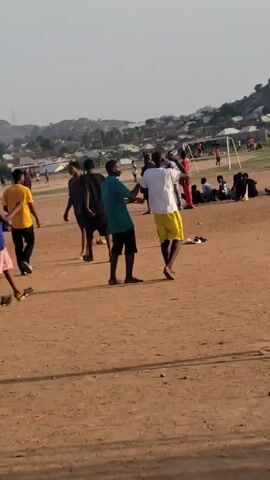 The video shows an outdoor dirt field populated with people, likely involved in or watching athletic activities. In the foreground, a young man wearing a purple t-shirt and dark shorts walks barefoot across the field. Other individuals are scattered across the field, some barefoot and others with shoes or sandals. A few people are sitting or standing in small groups. In the background, a soccer net is visible on the left side of the frame, with several people appearing to play soccer. A residential area with buildings and trees sits against a hilly backdrop in the distance. The lighting suggests it is either early morning or late afternoon.