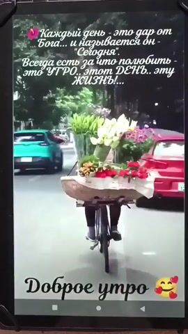 A person rides a bicycle loaded with a large bouquet of flowers. The person is wearing dark clothing. The bicycle is moving down a street lined with cars. The background is blurred, suggesting movement. Text overlays on the video read "Каждый день - это дар от Бога... и называется он "Сегодня". Всегда есть за что полюбить - это УТРО... этот ДЕНЬ.. эту ЖИЗНЬ!" which translates to "Every day is a gift from God... and it is called "Today." There is always something to love - this MORNING... this DAY.. this LIFE!" and "Доброе утро" which translates to "Good morning." There are also emojis of hearts and blushing faces.