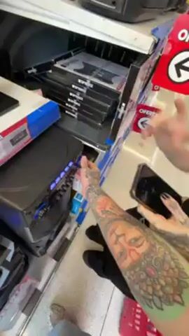A tattooed person holding a red shopping basket films a young woman with red hair and tattoos standing in an electronics aisle of a store. The woman looks at her phone and then examines items on a shelf filled with electronic devices. The aisle is also stocked with televisions. The filming person extends their tattooed arm out to either side of the frame. Music plays in the background.
