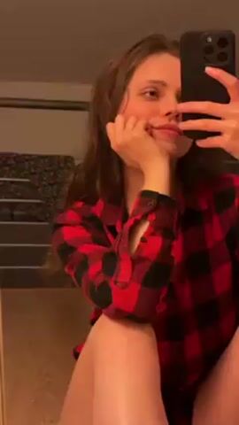 A woman is taking a selfie with a black iPhone. She is wearing a red and black checkered shirt. She is sitting with her legs visible, and her left hand is supporting her head. The background includes a bed, a wall, and some other furniture. The lighting is soft and warm.
