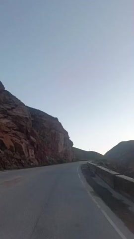 The video shows a view from a moving vehicle on a road that curves through a mountainous, arid landscape. A steep, red rock formation dominates the left side of the frame. On the right, a concrete barrier runs alongside the road, separating it from a darker, lower area. The sky is a pale blue, transitioning to a lighter hue near the horizon, suggesting either dawn or dusk. The road surface is asphalt, and there are no other vehicles or people visible.