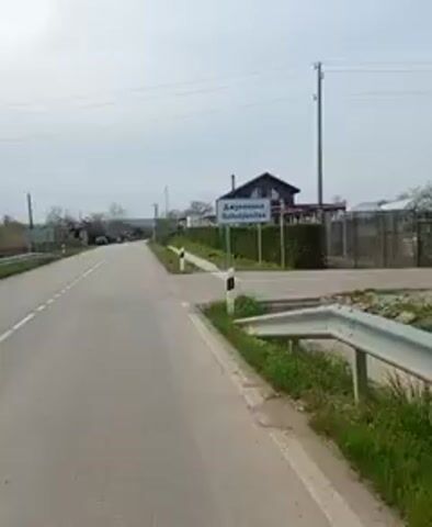 A person riding a blue and black mountain bike is passing by a sign that reads "Джулюница Dzhulyunitsa" in Cyrillic and Latin script. The road is paved and has a white line marking the edge. There is a building with a dark roof in the background, behind a green hedge and fence. The sky is overcast. The camera is positioned on the bike, providing a first-person perspective.