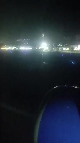 The video shows a view from an airplane window at night. The runway lights are visible in the distance, with blue lights marking the sides of the runway and brighter lights illuminating the area further away. A portion of the airplane's engine nacelle is visible in the foreground, colored blue and dark gray. The plane appears to be taxiing or preparing for takeoff or landing as evidenced by the runway lights.