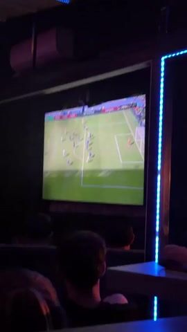 The video shows a large screen displaying a soccer game. The game is being played in a stadium with a green field and white lines. There are many players on the field, wearing blue and white uniforms. The camera is focused on the screen, and the environment around it is dark, suggesting it is nighttime or a dimly lit room. There are people in the foreground, their backs to the camera, watching the game. The screen shows a play developing near the goal, with players clustered around the net. One player in a red uniform, presumably the goalkeeper, is in the net. The screen also shows advertisements and logos, including "ARENASPORT" and "Lenovo." The overall scene suggests people are gathered to watch a soccer match.