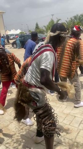 The video shows a group of people in traditional attire dancing outdoors. The main focus is on a man in a white t-shirt with red and black stripes over his shoulder, a cowrie shell belt, a fur skirt, and metal ornaments around his legs. He's holding a small, light-colored object in his hands, possibly a musical instrument. Surrounding him are other people, some wearing striped orange and brown shirts, red hats, and similar leg ornaments. They are all moving and dancing to the rhythm of the music.  Cars and buildings are visible in the background, suggesting an outdoor event or celebration. The ground is paved with concrete tiles.