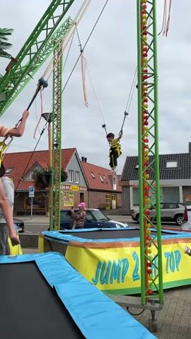 A child is bouncing on a bungee trampoline attraction. The child is attached to elastic cords that allow them to jump high into the air. A man in a gray sweatshirt is standing on the trampoline mat, looking at the child. There are two women with pink hats in the background, one of whom is looking at the camera. The attraction is set up in a street with buildings and parked cars in the background. The sky is cloudy.