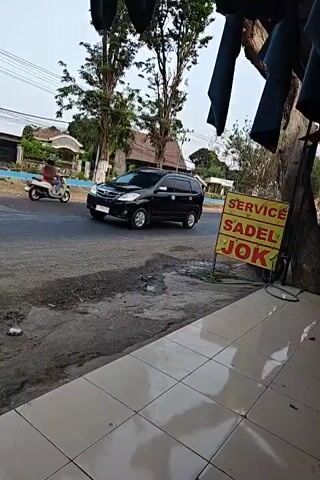 This video shows a roadside scene in what appears to be a rural or suburban area. On the left side of the frame, a black SUV is parked or moving slowly on a paved road. Further down the road, a motorcycle with a rider is visible. On the right side of the frame, a yellow sign with red and black lettering reads "SERVICE SADEL JOK," indicating a service for saddle or upholstery repair. The sign is propped up on a grassy area next to a tree and a tiled sidewalk. In the background, there are trees, houses, and power lines under an overcast sky. The foreground is dominated by the tiled sidewalk. The video is taken from a slightly elevated, static perspective.