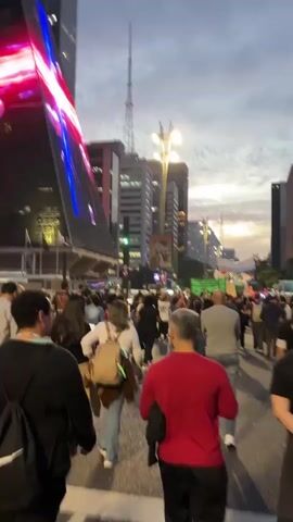 The video depicts a large group of people walking down a city street at dusk. The crowd consists of diverse individuals, dressed in various casual clothing. There are multiple tall buildings lining the street, with one notable structure on the left displaying bright, colorful lights on its facade. Street lamps are visible, and the sky is overcast, with the setting sun creating a warm glow on the horizon. The movement of the camera suggests the video is taken from the perspective of someone walking with the crowd, providing a close-up view of the people and their surroundings. The overall atmosphere is busy and dynamic, capturing the energy of a large gathering in an urban environment.