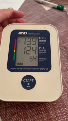 The video shows a blood pressure reading being taken on a person's arm. The blood pressure machine is white with a blue display. The reading is 139/124 with a pulse of 54. The person is speaking in Russian, but it's not possible to hear what is said. The blood pressure cuff is on the person's left arm. The background is a burgundy woven surface.