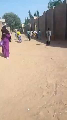 The video shows a group of children running down a sandy road in a village or town. Most of the children are wearing brightly colored clothing, including yellow, green, and blue. One child is wearing a black outfit. A person in a long purple dress is walking in the opposite direction. Buildings with high walls line both sides of the road. The sky is blue and clear, indicating a sunny day.  The road surface is uneven with visible footprints and scattered debris.