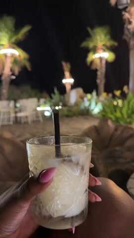 The video shows a first-person perspective of someone holding a clear glass with a light-colored drink and a black straw. The drink contains ice and appears to have some garnish. The person's hand, with pink-painted fingernails, is visible holding the glass. The background is a nighttime outdoor setting, likely a beach or patio. Several palm trees are illuminated by string lights wrapped around their trunks. In the mid-ground, there are tables and chairs, some with white tablecloths, and lush green foliage. In the far background, there is a dark sky. The scene suggests a relaxed atmosphere, possibly a restaurant or bar. The camera is stationary and the video consists of a series of still frames.