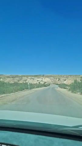 The video shows a view from inside a vehicle traveling on a paved road through a desert landscape. The road curves to the left, leading towards a distant horizon of rolling hills and sparse vegetation under a clear blue sky. The terrain on either side of the road is dry and sandy, with scattered green bushes and some light-colored, eroded hills. In the distance, a few small structures or vehicles can be seen on top of the hills. The vehicle's dashboard is visible at the bottom of the frame. The overall impression is one of a solitary journey through a vast, arid environment.