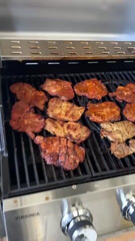 The video shows several pieces of meat grilling on a gas grill. The pieces of meat vary in color from reddish to browned, and some have visible grill marks. The grill is stainless steel with a black interior and black cooking grates. The knobs for the grill are located on the lower right side of the grill. The brand name "VIRGINIA" is visible near the knobs. The camera moves slightly around the grill to show the different pieces of meat cooking.