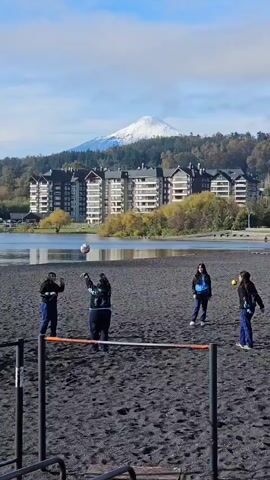 The video shows a group of people playing on a black sand beach. There are four people initially visible near a low horizontal bar structure, passing a ball back and forth. Another person joins the group and kicks the ball.  In the background, there is a calm lake with a mountain partially covered with snow. Several multi-story buildings line the edge of the lake. The sky is blue with scattered clouds.