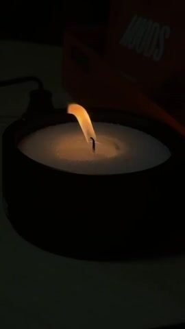 A close-up shot of a candle with a lit flame. The candle is in a black bowl filled with white material. In the background, a red box with white lettering that reads "MOODS" is visible. The lighting is dim, with the flame providing the main source of light. The flame is golden yellow and flickers gently. The candle wick is black and stands upright. The white material around the wick appears to be granular. The overall mood is calm and serene.