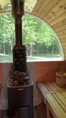 The video is taken from inside a wooden structure that appears to be a sauna. In the foreground, a black sauna stove with stones on top is visible. A metal chimney extends upwards from the stove. Through a large, arched window, a sunny outdoor scene is visible, featuring a green lawn and numerous trees with lush foliage. The camera pans slightly to the right, revealing a wooden bench inside the structure and a small wooden bucket with ladle on it.