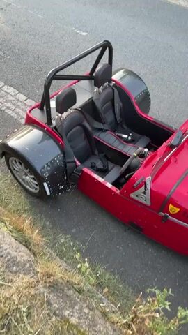 A red, three-wheeled vehicle is parked on the side of a road. It has two seats, a roll bar, and a license plate that reads "TI-GER". The car is partially on the curb, with some grass and moss visible nearby. The road is paved and there is a sidewalk on the other side. The weather appears sunny.