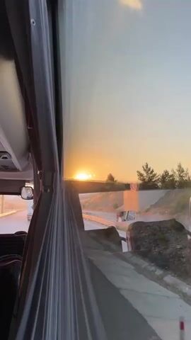 The video begins from inside a vehicle, likely a bus, looking out the window. The interior of the bus is visible with seats and curtains. Outside, the sun is setting, casting a warm glow over the sky. The camera pans to capture the scenery, which includes a highway overpass, some greenery, and a hazy sky with the sun partially obscured by clouds. The sun appears as a bright orb, and a reflection of it is also visible in the sky, creating a lens flare effect. The video briefly shows the blurred interior of the vehicle. The overall scene depicts a sunset viewed from a moving vehicle on a highway.