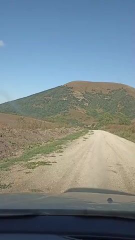 The video shows a view from inside a vehicle driving on a dirt road. The road is unpaved and appears to be in a rural or natural setting. To the left of the road, there are fields, some of which appear to be tilled or cultivated, and some with sparse vegetation. Further in the distance, a large, rounded hill or mountain covered in vegetation dominates the landscape. The sky is clear and blue with a few wispy clouds visible. The vehicle is moving forward, indicated by the shifting perspective and the road stretching ahead. The overall impression is of a scenic drive through a natural, undeveloped area.