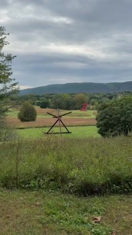 The video displays a wide shot of a rural landscape under a cloudy sky. In the foreground, tall green grass and wildflowers are visible. In the middle ground, a large, dark, triangular sculpture stands in a grassy field. Further in the distance, a bright red abstract sculpture resembling a stylized figure stands on a small hill. The background features rolling green hills and trees, with a larger mountain range in the far distance. The overall impression is one of an outdoor art installation in a natural setting.