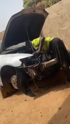 The video shows a man working on a car engine. The car is white and appears to be quite old and damaged, with missing parts and rust. The hood is open, and the man is leaning over the engine compartment, seemingly working on the engine. He is wearing a bright yellow shirt and dark pants. The car is propped up on cinder blocks. The background consists of a brick wall and a few trees. The setting suggests a somewhat outdoor or semi-enclosed workspace.