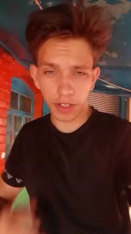 A young man with short, messy brown hair and blue eyes, wearing a black t-shirt, looks directly at the camera. His mouth is slightly open as if he is speaking. He is standing in what appears to be an outdoor setting with a blue and red structure behind him, possibly a porch or a covered area. There is a white string with a light bulb hanging from the ceiling structure in the background. The man's right hand is visible, with his fingers slightly curled. He wears a black watch on his left wrist.