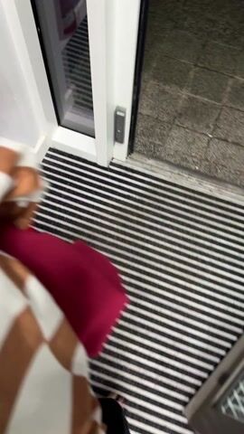 The video appears to be shot from a first-person perspective. A person is walking outside on a paved area, possibly a sidewalk or walkway. The person is wearing flip-flops, black pants, and a shirt with brown and white stripes. They are carrying a red item. The ground is made of rectangular pavers. To the side, there is a patch of green grass and a building with green and white accents, possibly a laundry room or similar service area. The lighting indicates it is either nighttime or a very overcast day. The camera movement suggests the person is walking at a normal pace.
