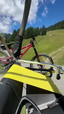 The video shows a person riding a chairlift up a mountain on a sunny day. There is a red mountain bike attached to the chairlift in front of them. The mountainside is covered in green grass and trees, and there is a dirt path winding up the hill. Other people are walking and biking along the path. The chairlift passes over a small wooden structure.