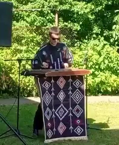 A man wearing a black shirt with colorful patterns and sunglasses stands behind a wooden instrument on a table. The table is covered with a dark fabric with diamond-shaped patterns. The man is holding two mallets and appears to be playing the instrument. He is outdoors in front of a fence and trees. A speaker is visible to the left.