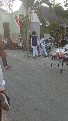 The video shows a group of people, mostly children, in what appears to be a procession or event. The camera is held at a low angle, and the footage is oriented vertically.

In the foreground, a person's lower legs and shoes are visible, suggesting the camera is being held by someone walking or standing.

The main focus of the video is a line of individuals, predominantly children, walking on a paved surface. Many of the children are wearing uniforms, including white shirts and either brown or grey vests. Some of the girls are wearing headscarves. One child in the foreground, to the right, is wearing an orange shirt and a white cap, and appears to be looking up and pointing.

Behind the children, there are adults, some of whom are also wearing headscarves or caps. A building, likely a school or religious institution, is visible in the background, with a green facade and a white trim. An Indonesian flag is also visible, partially obscured by trees.

There is a palm tree to the left of the frame. The sun is shining brightly, casting long shadows and creating a lens flare effect. There are some parked motorcycles in the background near the building. The overall scene suggests an outdoor event taking place during daylight hours.
