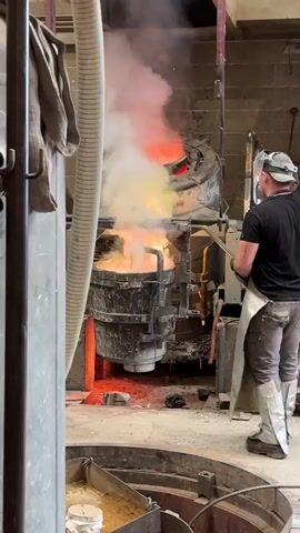 A worker stands in a metal foundry, observing a crucible filled with molten metal. The crucible is positioned beneath a furnace, with flames and smoke rising from both. Sparks fly as the worker stands by a steel basin. The worker wears protective gear, including a visor and apron. Industrial equipment and hoses are visible around the scene. The overall atmosphere is intense, with the heat and light from the molten metal dominating the setting.