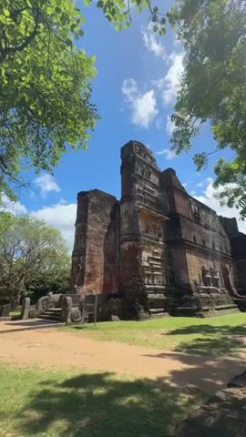 The video shows an ancient brick structure surrounded by greenery and trees. The sky is blue with scattered white clouds. The structure appears to be a ruin or a historical building, showing signs of age and wear. The surrounding area includes a lawn and dirt paths. The camera pans slightly from right to left, revealing more of the surrounding trees and greenery.